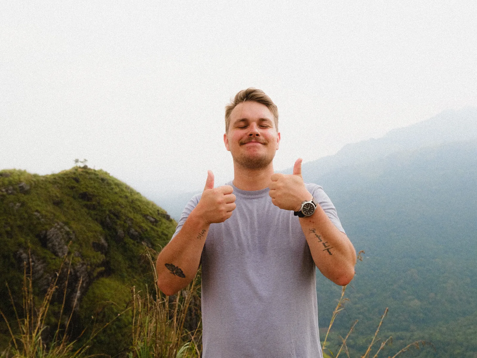 Dennis giving two thumbs up on a mountain, mist and green hills behind him